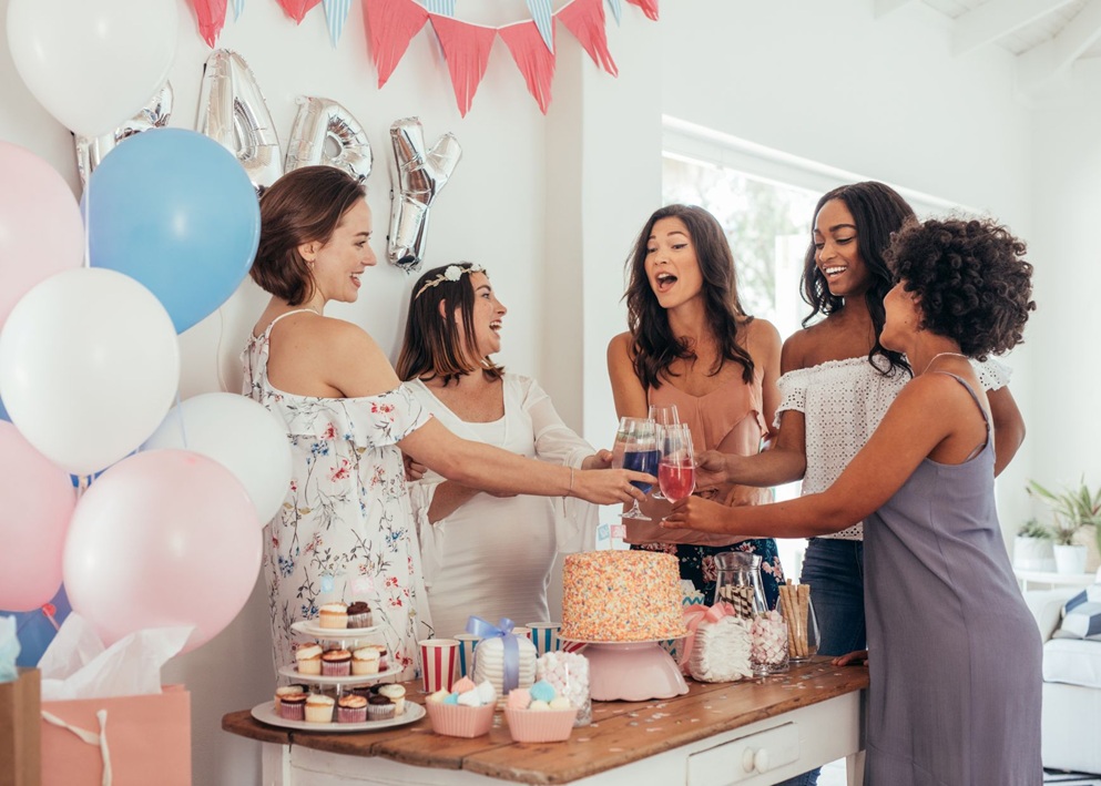 Five women raising glasses at baby shower table with cake, cupcakes, balloons, and festive bunting.