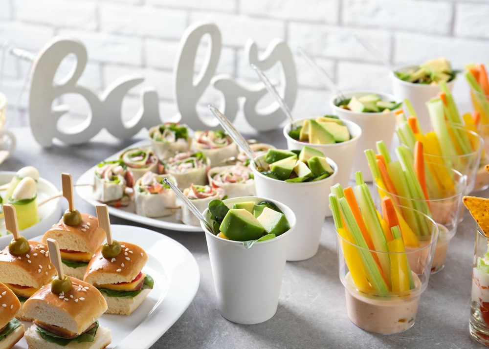 Appetizer table with mini sandwiches, tortilla roll-ups, avocado cups, and vegetable sticks with dip.