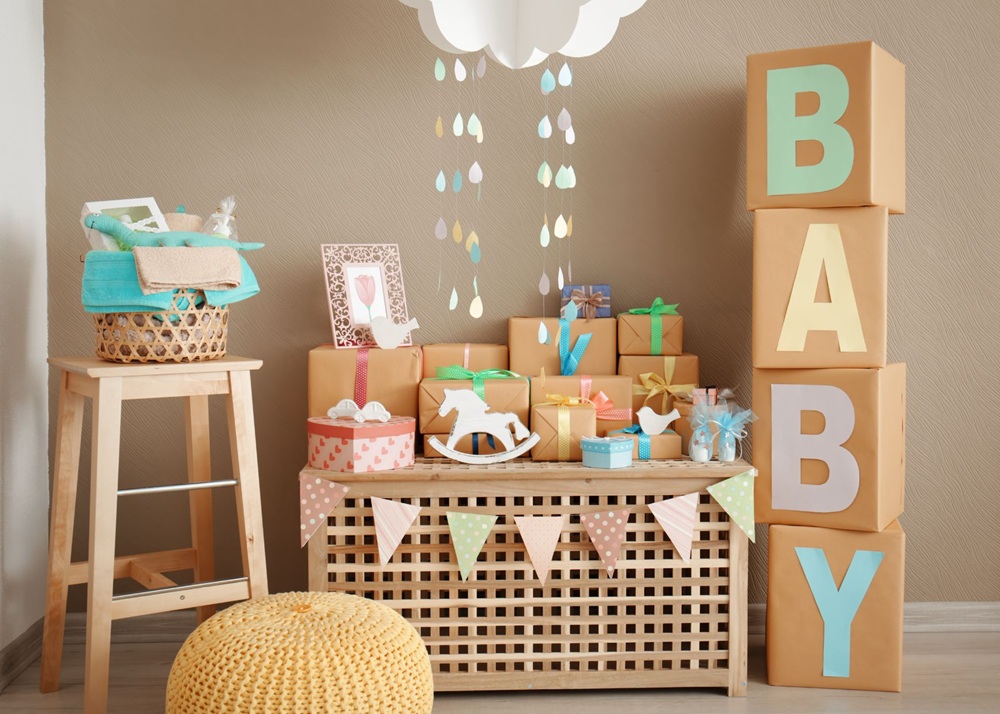 Pastel baby shower decorations with gift boxes, rocking horse, and cloud mobile above wooden chest.