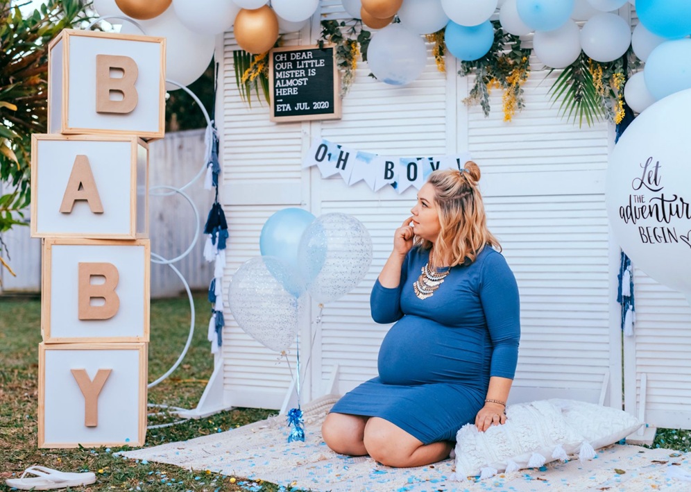 Pregnant woman seated outdoors with blue balloons, BABY blocks, OH BOY banner, and adventure balloon.