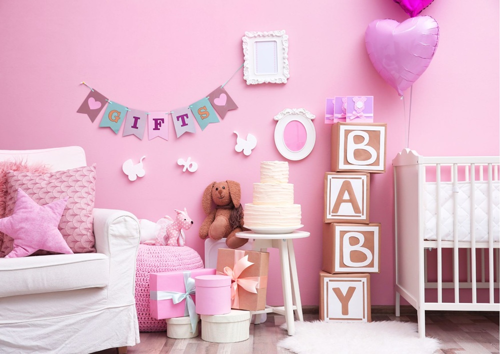 Pink-themed baby shower setup with cake table, stuffed toys, gift boxes, crib, and decorative banner.