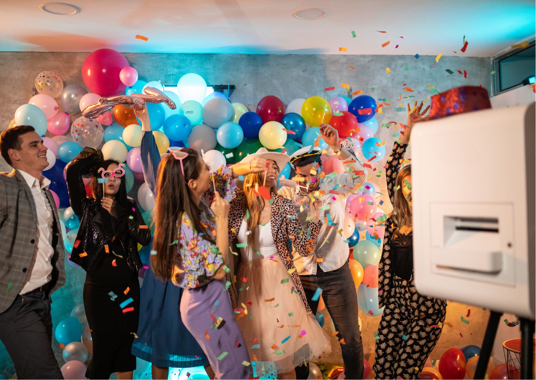 Group celebrating with confetti, vibrant outfits, and multicolored balloon wall backdrop.