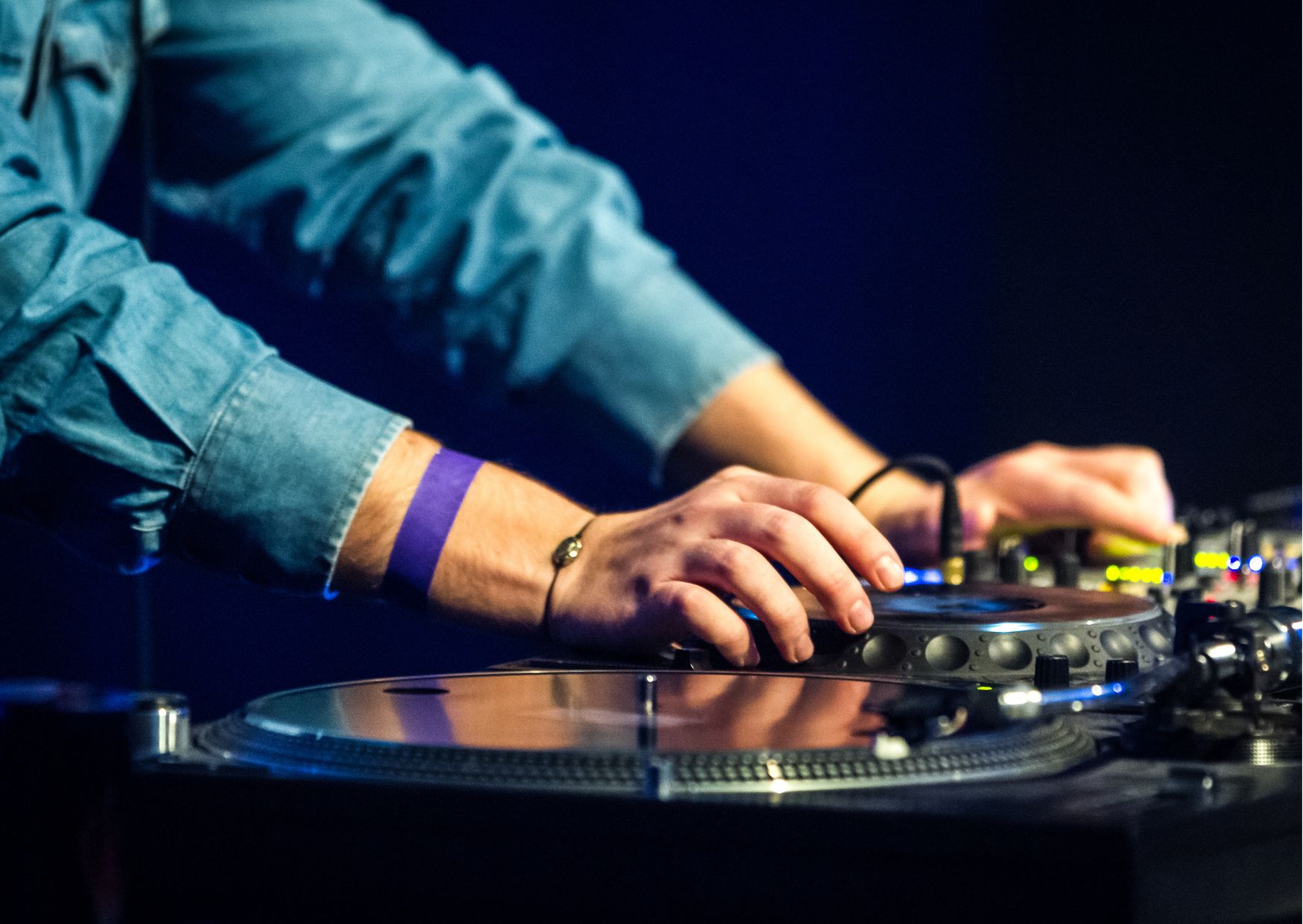 Close-up of DJ operating turntable and mixer, wearing denim shirt and wristband, illuminated by stage lighting.