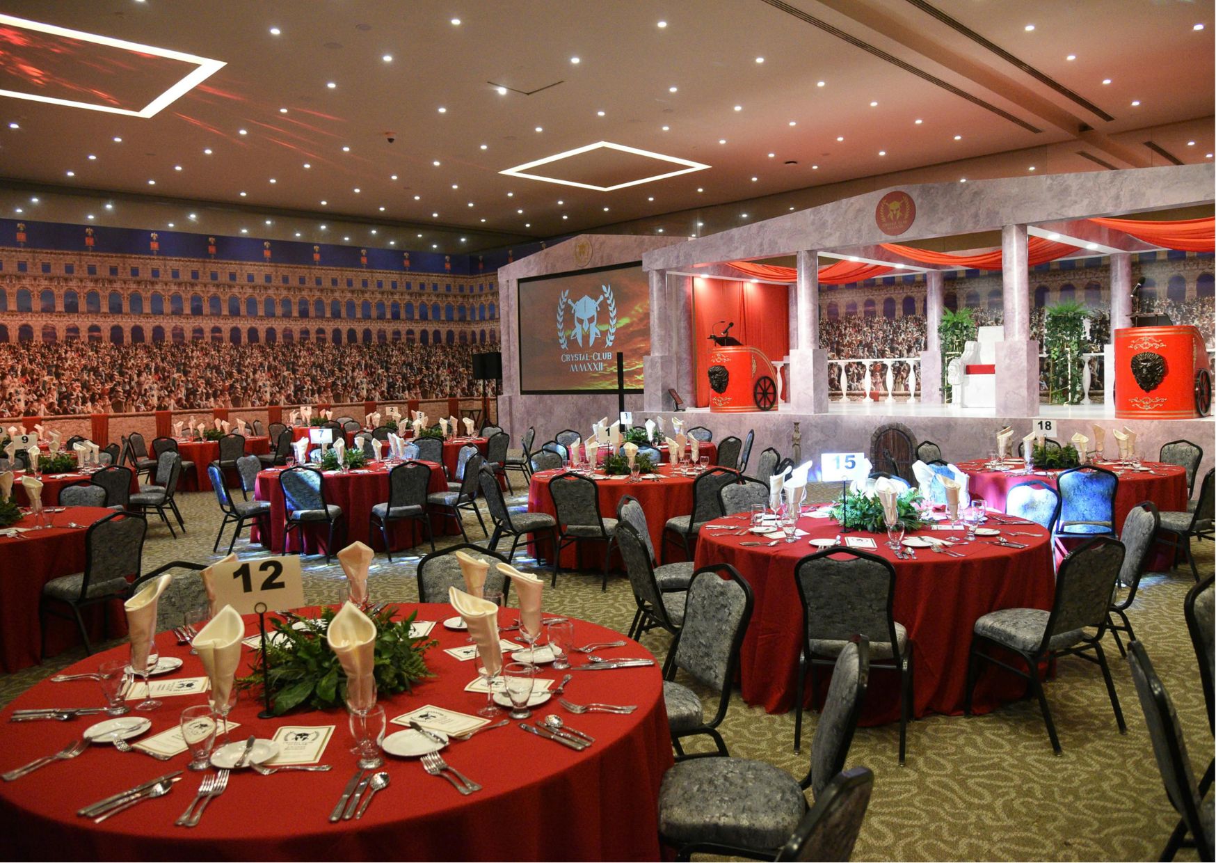  Red-draped stage with Roman columns, themed murals, and formal dining tables.