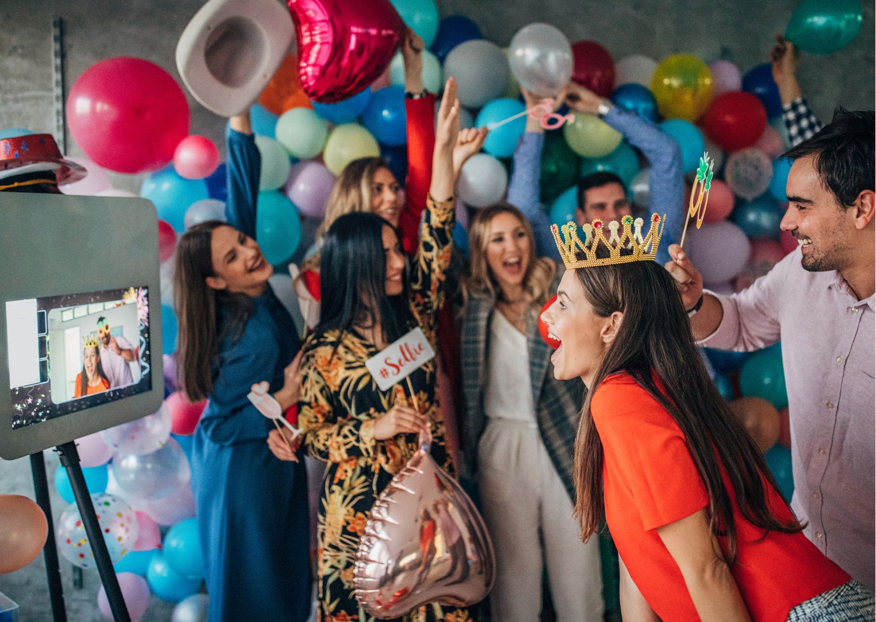 Group of people posing with props in front of a colorful balloon backdrop at a lively party photo booth.