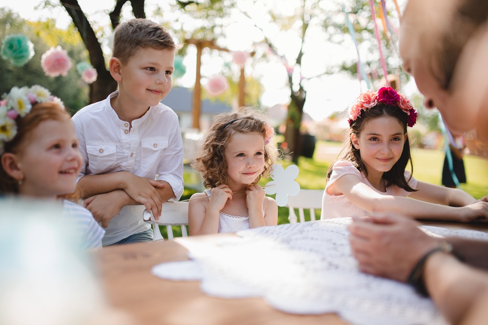 Children enjoying outdoor garden party seated at table with colorful decorations and festive atmosphere in background.