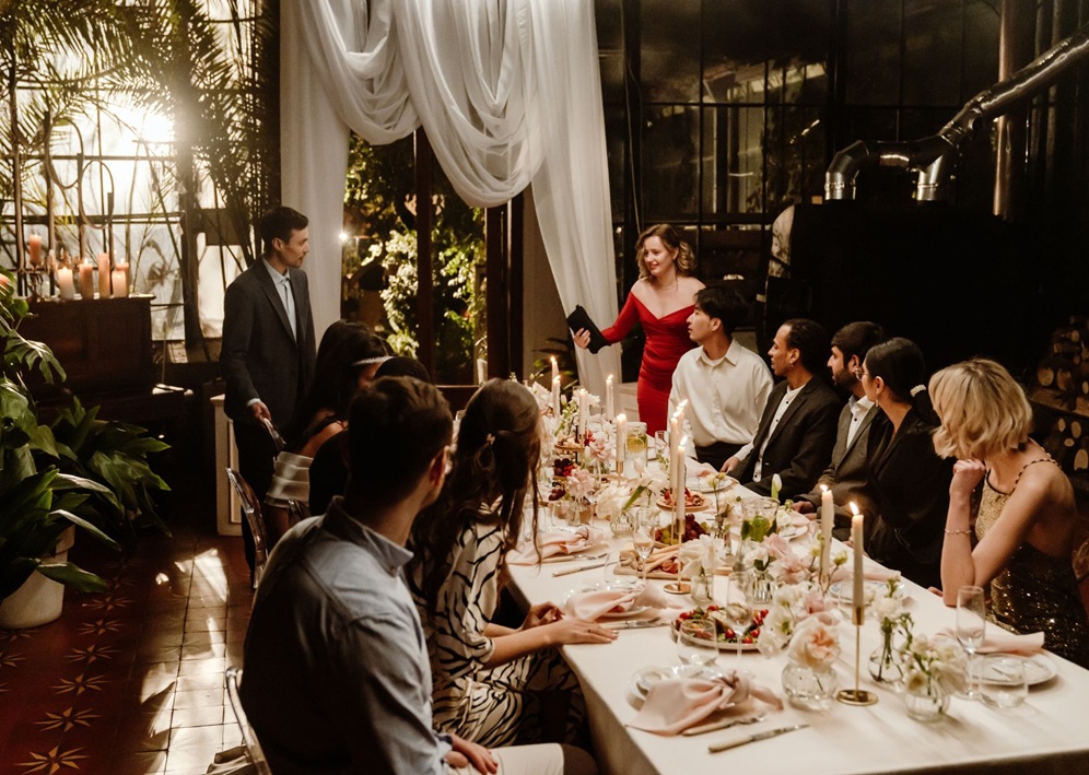 Elegant dinner party with guests seated at long table with candles, flowers, and draped white fabric backdrop