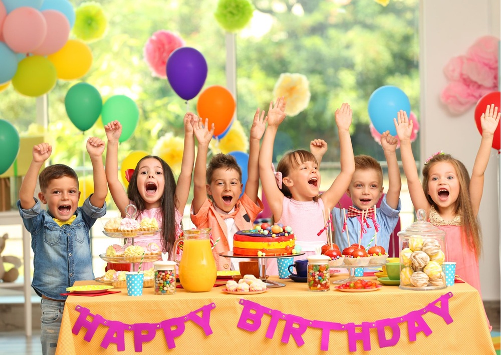 Excited children celebrating at birthday party table with colorful balloons, cake, and Happy Birthday banner