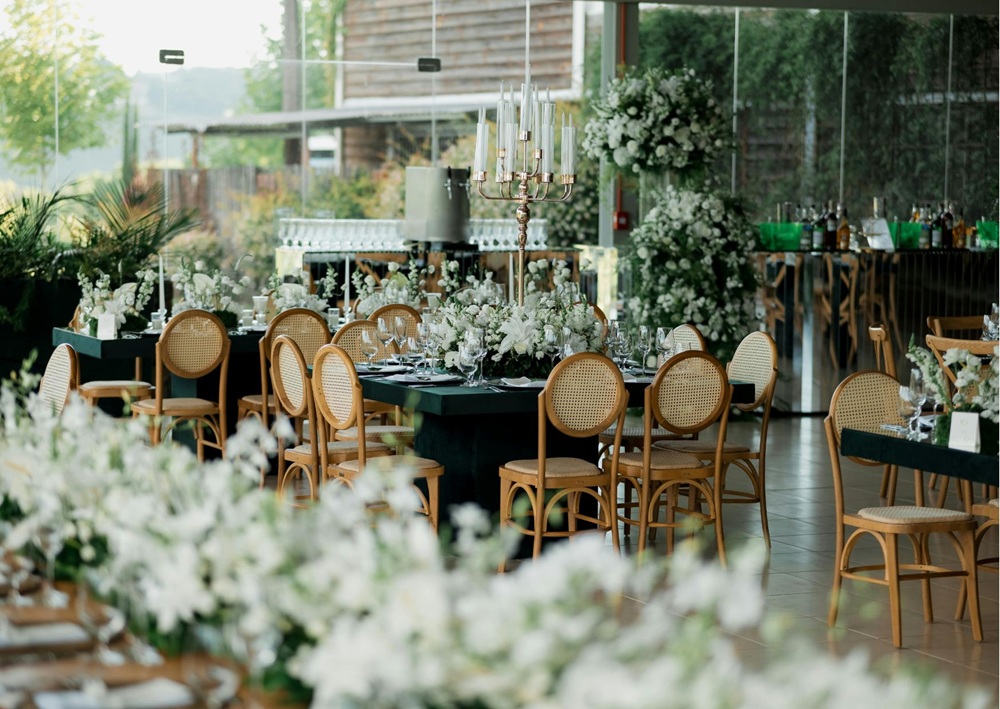 Contemporary wedding reception with black tables, rattan chairs, white floral arrangements, and glass walls overlooking greenery