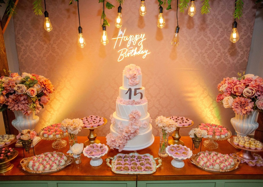 Elegant fifteenth birthday celebration with tiered cake, Happy Birthday neon sign, hanging Edison bulbs, and pink floral arrangements