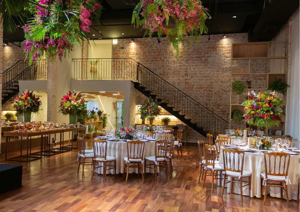 Reception venue with round tables, hanging tropical floral arrangements, exposed brick walls, and warm ambient lighting