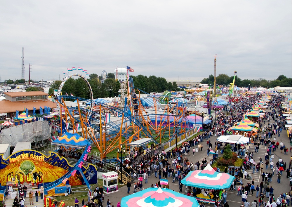 Aerial view of outdoor carnival with colorful rides, tents, and crowds at fairground.