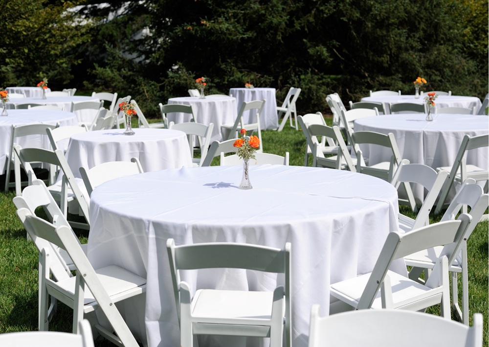 White folding chairs and round tables with linens arranged on green lawn for outdoor event.