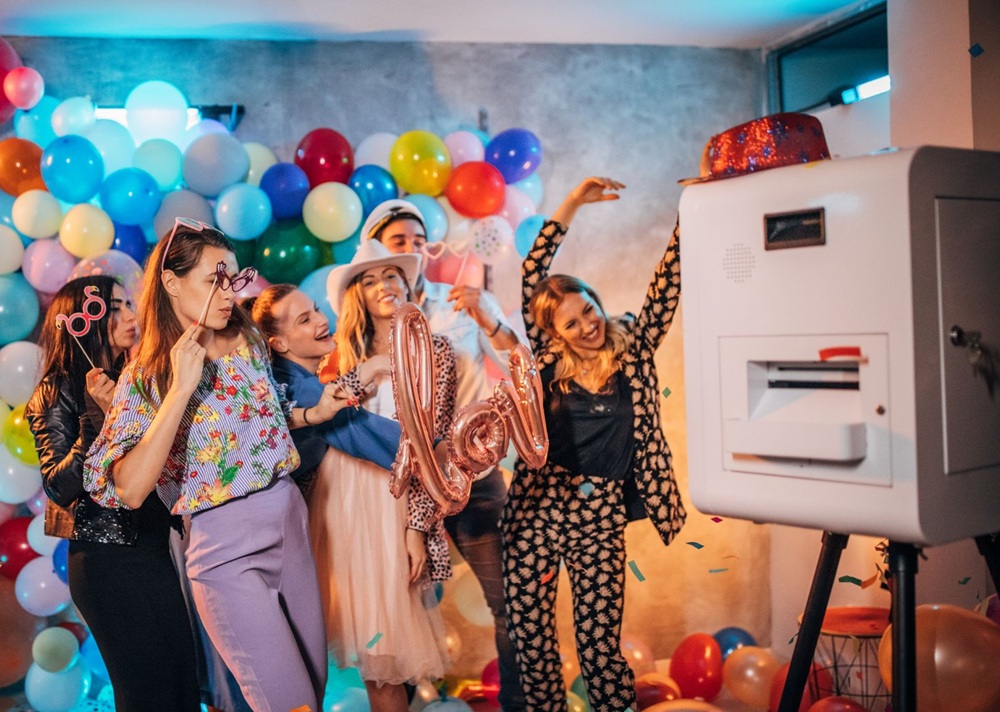 Friends dance and pose beside a photo booth surrounded by balloons and confetti.