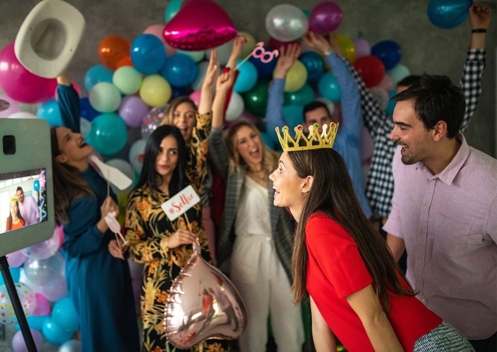 A lively group celebrates in front of a photo booth with balloons and props.