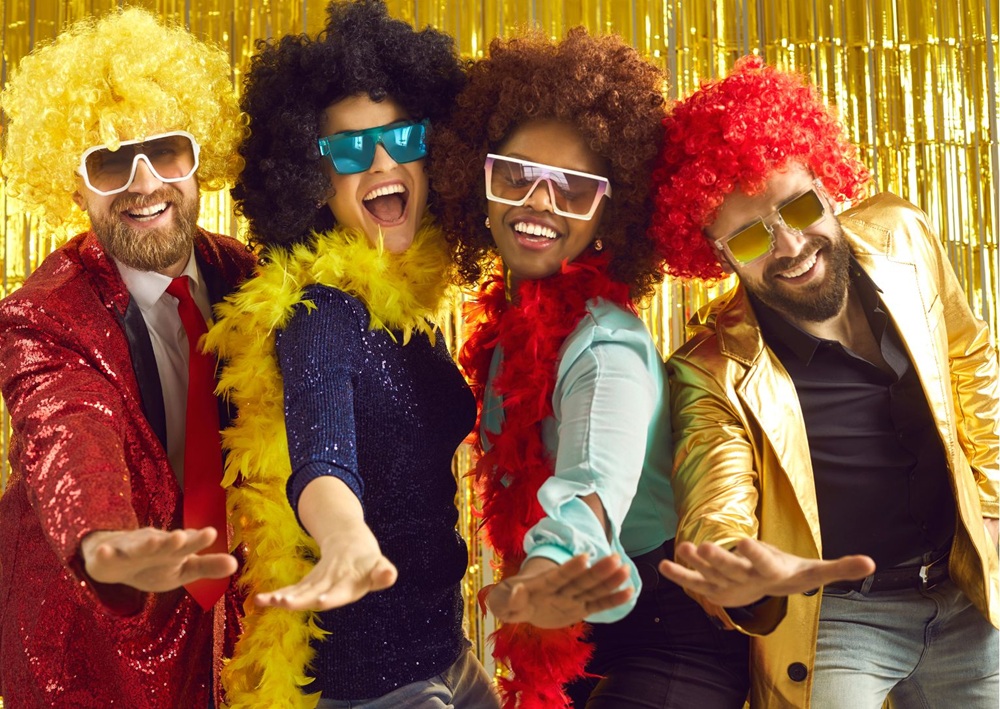 Four friends in colorful wigs and glasses pose joyfully at a festive party.