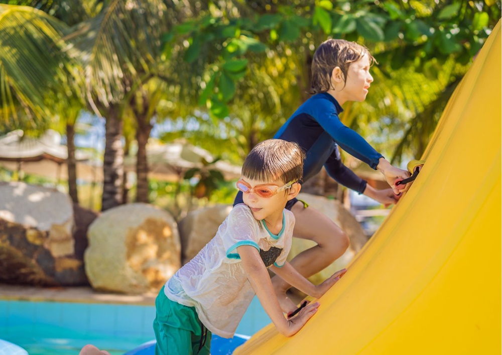 Children climbing a tall inflatable slide beside palm trees and a pool.