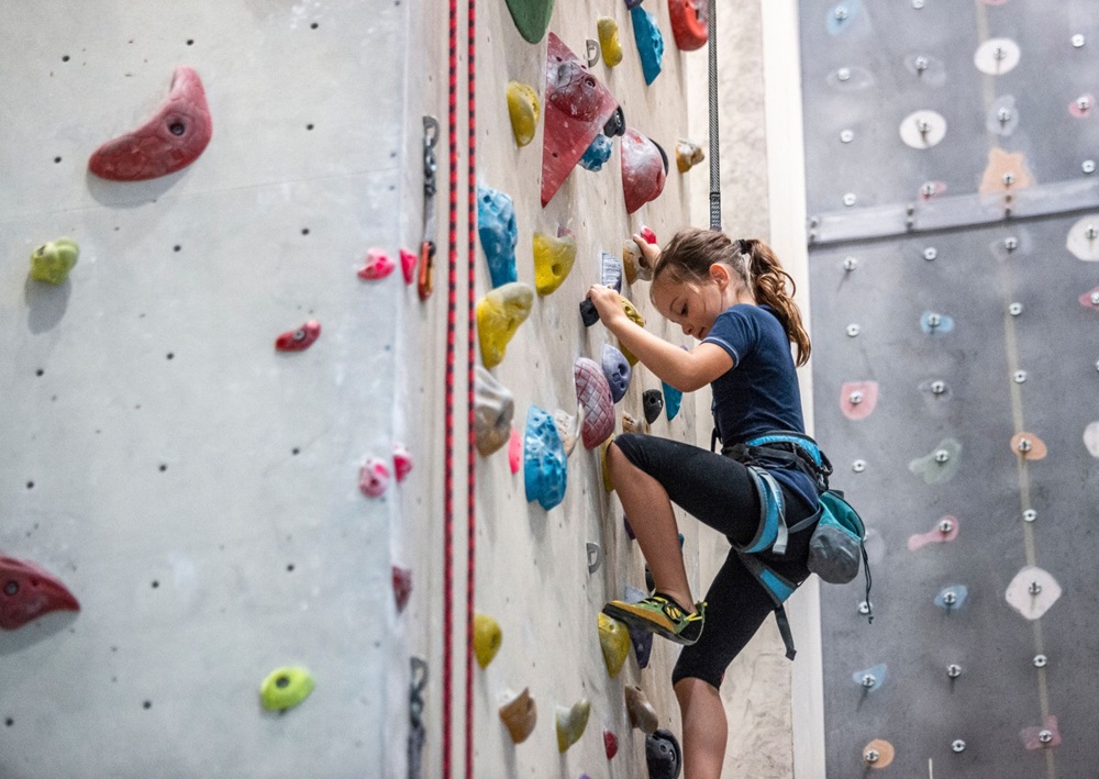 Young climber scales an indoor rock wall using colorful holds and safety gear.